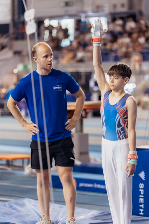 Young male gymnast raises hand to salute before his routine as his coach watches attentively in a packed arena.