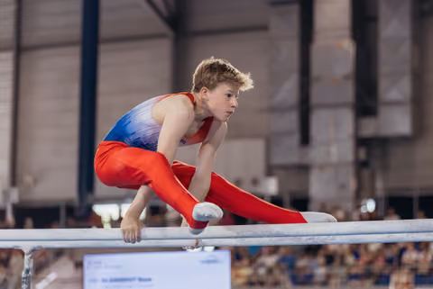 Young male gymnast performs a straddle hold on parallel bars, his focused expression intense during a competition.