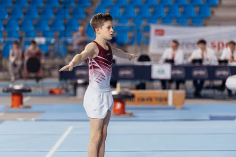 Young male gymnast performs on floor, arms outstretched, focused expression, in a blue-matted arena with judges behind.