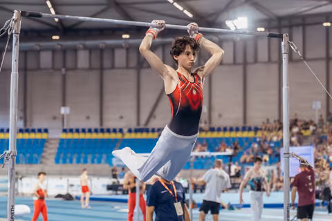 Young male gymnast hangs from the high bar with focused intensity, wearing red and black leo, in a large sports hall.