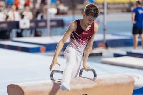 Young male gymnast concentrates intensely while performing on the pommel horse, wearing a red Quatro leotard.