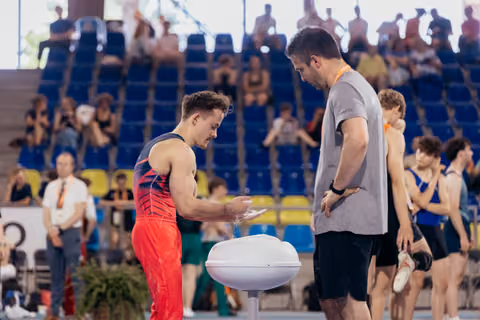 Young gymnast applies chalk to hands at a bowl while his coach watches attentively in a busy competition arena.