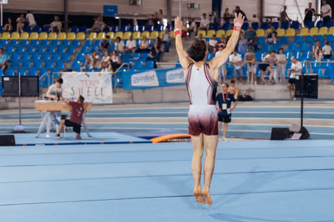Male gymnast raises both arms triumphantly on the floor exercise mat, back to camera, crowd watching in the arena.