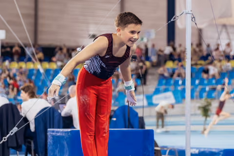 Young male gymnast smiling proudly on parallel bars, wearing red pants and grips, in a packed indoor arena.