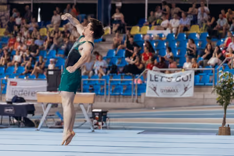 Male gymnast leaps with fist raised in triumph, green uniform, performing before a packed indoor arena crowd.