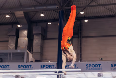 Male gymnast performs a perfect handstand on parallel bars, body fully extended upward, wearing red Belgian uniform.