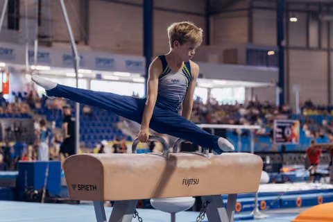 Focused young male gymnast executes a scissor split on a Spieth pommel horse during a competitive gymnastics event.