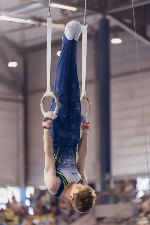 Young male gymnast performs an inverted hang on still rings, legs extended upward, showing intense concentration in a competition hall.