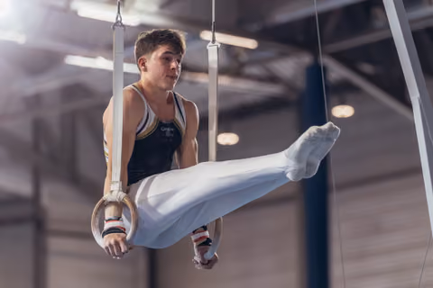 Young male gymnast performs on still rings with intense focus, legs raised, wearing white pants and dark vest in a gymnasium.