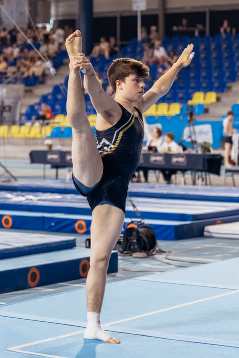 Young male gymnast performs a standing leg hold on the floor, focused expression, in a blue gymnastics arena.