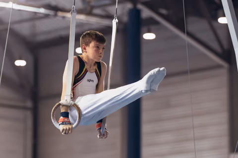Focused young male gymnast holds still rings with legs raised, wearing agivo kit in a competition gymnasium.