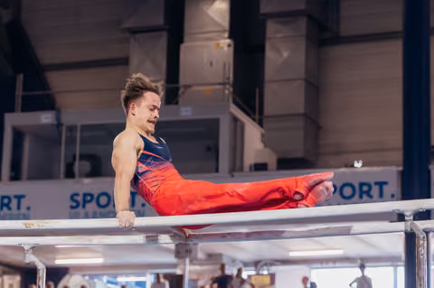 Male gymnast performs a straddle L-sit on parallel bars, jaw clenched in intense concentration, at an indoor sports hall.