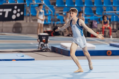 Young male gymnast performs floor routine, arms extended with focused concentration in a blue arena setting.