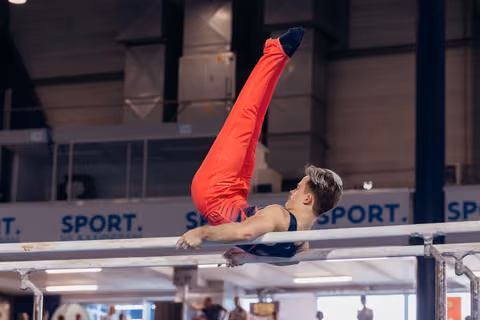 Young male gymnast performs a straddle-L hold on parallel bars, legs raised high, focused expression in a sports hall.