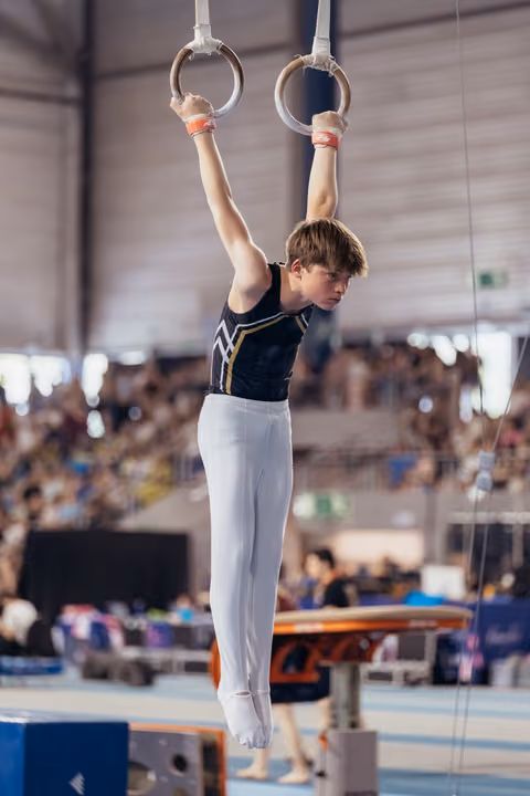 Young male gymnast hangs from still rings with intense focus, arms extended overhead, wearing black and white leotard.