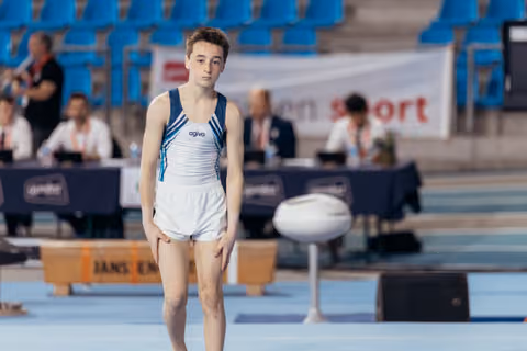 Young male gymnast stands still on the floor, focused and composed, wearing an Agiva leotard at an indoor gymnastics meet.