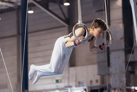Young male gymnast performs on still rings, body horizontal, face set with fierce concentration in an indoor sports hall.