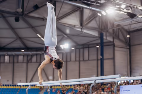 Male gymnast executing a precise handstand on parallel bars, legs perfectly vertical, in a packed sports arena.