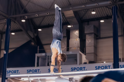 Male gymnast performs a perfect handstand on parallel bars, body fully extended, in an indoor sports hall.