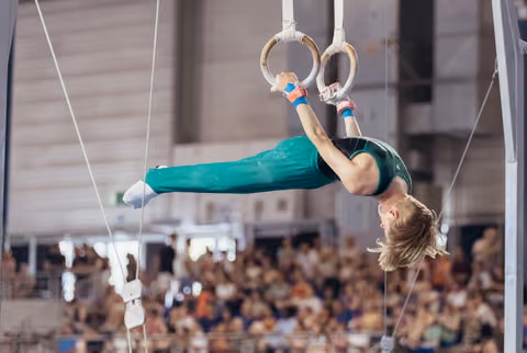Young male gymnast performs an inverted hold on still rings, body fully extended, intense focus, before a packed crowd.