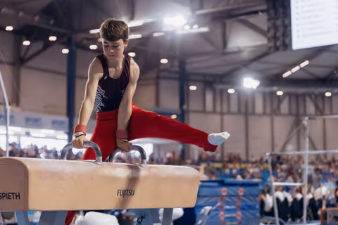 Young male gymnast performs focused pommel horse routine, leg extended, in a large indoor arena with spectators.