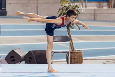 Young male gymnast performs a precise arabesque balance on floor, arms outstretched, expression intense and focused.