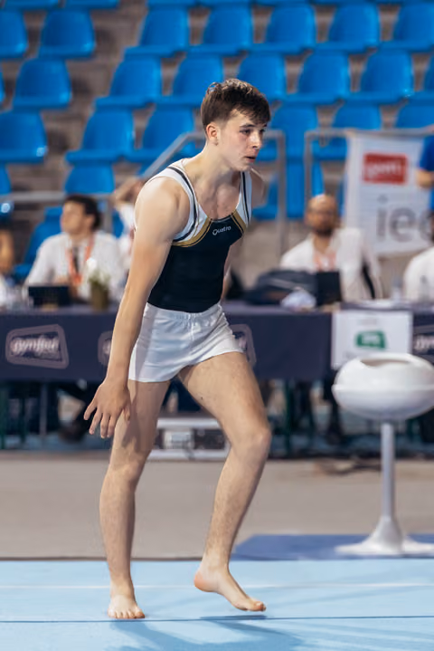 Young male gymnast in focused crouch position on floor exercise mat, wearing black Quatro leotard, in a competitive arena.