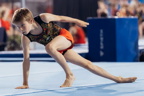 Young gymnast performs a focused floor routine lunge, arms extended, showing intense concentration during competition.
