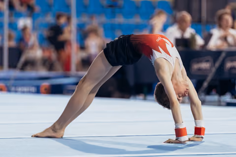Male gymnast performing a back bridge on the floor exercise mat, wearing a red and black leotard at a competition.