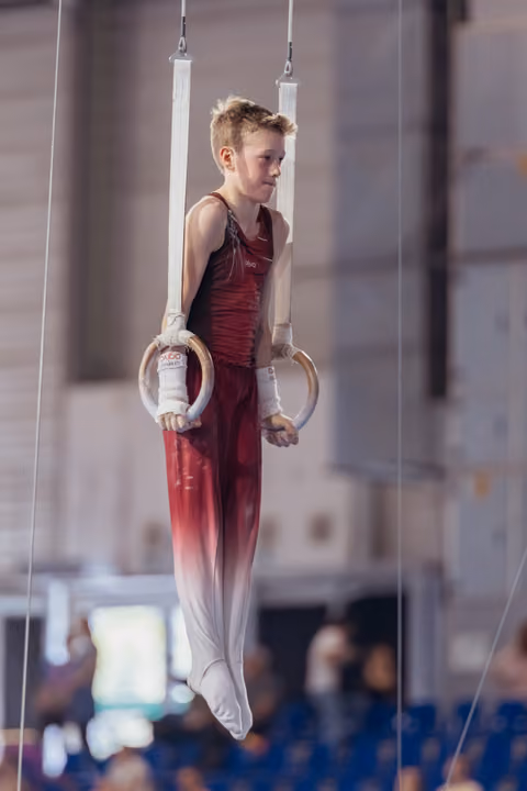 Young male gymnast in red uniform hangs still on rings, focused expression, competing in indoor gymnastics hall.