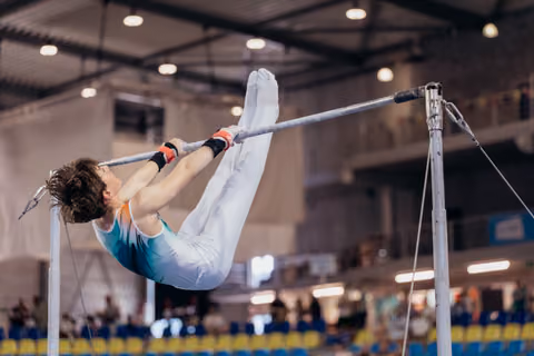 Young male gymnast performs a tuck on the horizontal bar, body curved with intense focus, inside a bright competition hall.
