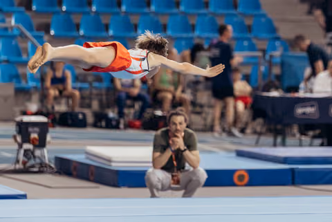 Young gymnast soars in a superman dive over the floor mat while a focused coach crouches and watches intently below.
