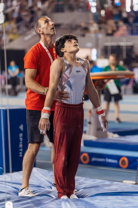 Young male gymnast and coach both gaze upward, coach steadying him with hands on shoulders in a supportive moment.