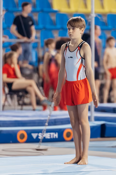 Young gymnast stands barefoot on the mat, composed and focused, wearing white and red leotard with shorts, awaiting their turn.