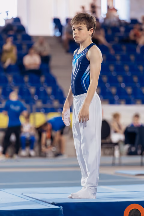 Young male gymnast stands still on the floor mat, focused and composed, wearing a blue leotard and white trousers.