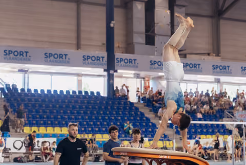 Male gymnast performs handstand on vault while coaches watch attentively in a Sport Vlaanderen arena.