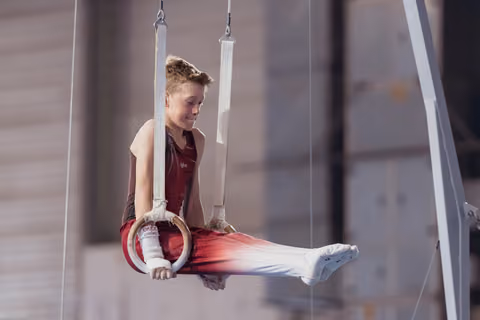 Young male gymnast performs on still rings, legs extended, showing concentration and control in a gymnasium setting.
