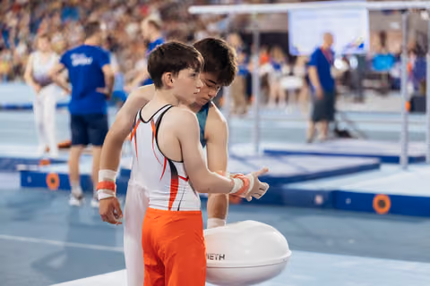 Young male gymnast gets wrist support from coach at chalk bowl, both focused before apparatus routine.