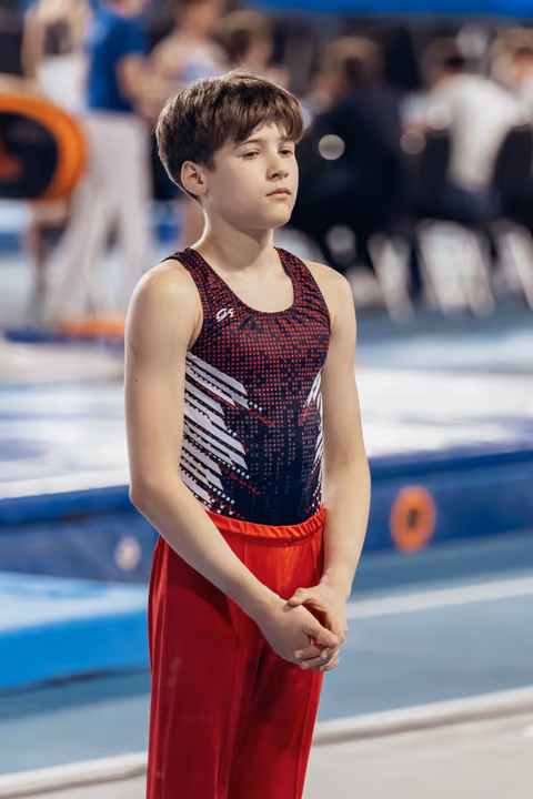 Young male gymnast stands composed, hands clasped, with a calm focused expression before his floor routine.