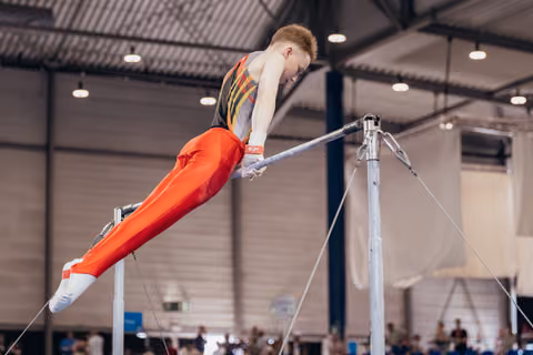 Young male gymnast swings powerfully on the horizontal bar, body extended, focused expression in an indoor sports hall.