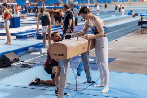 Coach kneels to adjust a Spieth pommel horse while a young gymnast in a leotard watches attentively in a busy arena.
