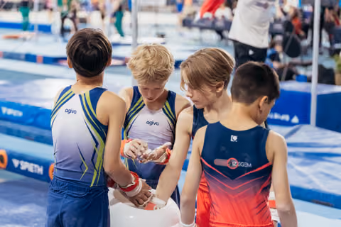 Four young male gymnasts huddle together applying chalk to their hands, focused and preparing before their routine at a gymnastics meet.