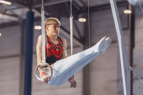 Young male gymnast holds still rings with intense focus, legs raised in pike position, wearing red and black leotard.