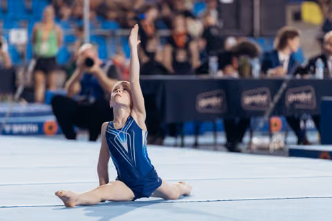 Young gymnast performs splits on floor, one arm raised skyward, arching back with focused expression during competition.