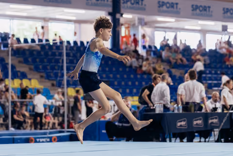 Young male gymnast leaps powerfully across the floor exercise mat, focused and composed at an indoor gymnastics meet.