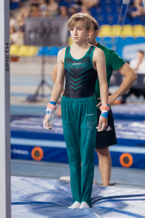 Young male gymnast stands composed at the floor, chalked hands ready, wearing teal Quatro uniform in a competitive arena.