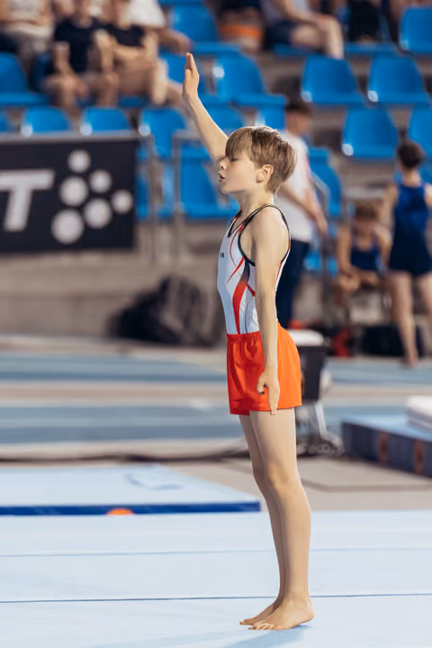 Young male gymnast stands barefoot on floor mat, raising one hand to salute the judges before his routine.