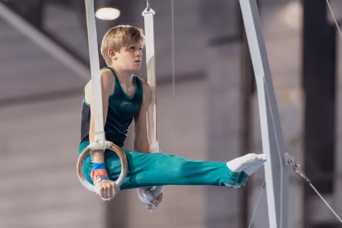 Young male gymnast holds a still rings position with intense focus, legs extended, wearing teal uniform in gymnasium.
