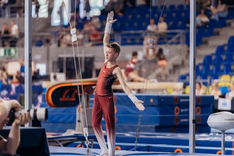 Young male gymnast raises one hand in a confident salute on the trampoline, wearing red suit and chalk-covered grips.
