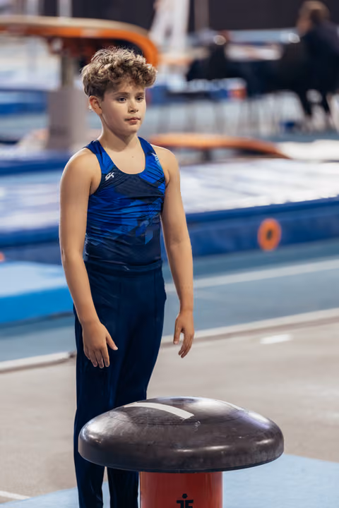 Young male gymnast stands focused beside the vault, wearing blue GK leotard, ready to compete in a gymnasium.